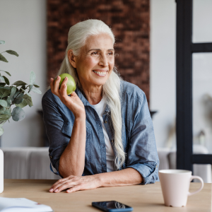 A smiling elderly woman with long white hair, wearing a denim shirt, sits at a table holding a green apple. She is looking off to the side, appearing relaxed and content. On the table in front of her is a white mug, a smartphone, and some papers.
