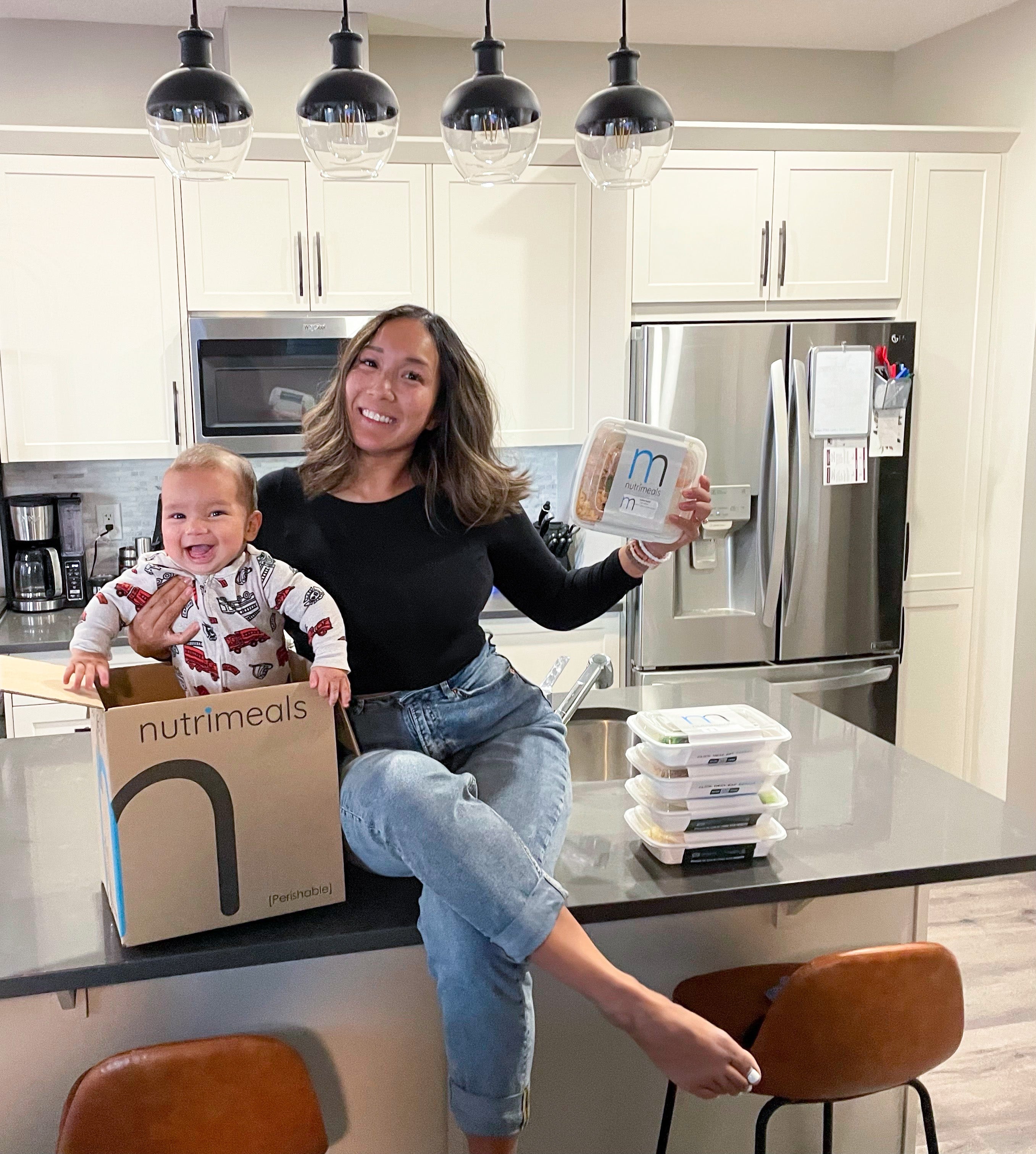 mom sitting on counter top with baby holding a meal prep container