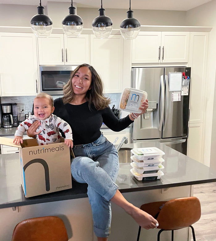 mom sitting on counter top with baby holding a meal prep container